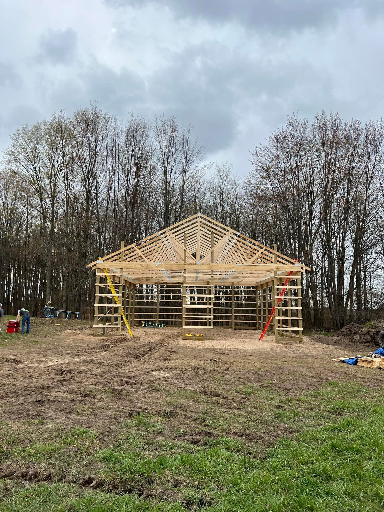 Wooden frame of a building under construction in a field, with trees in the background and a cloudy sky.