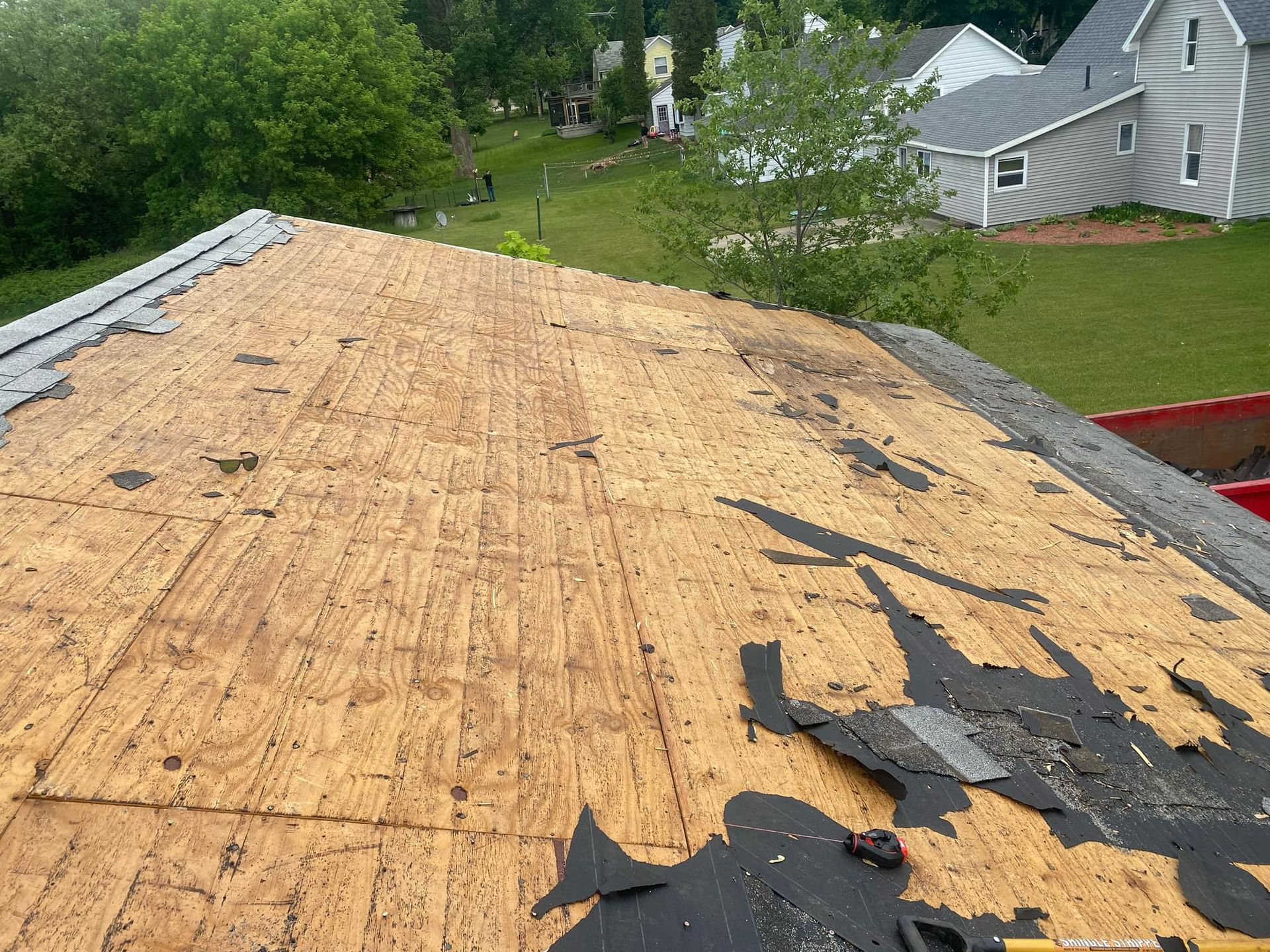 Roof with missing shingles revealing wood sheathing and a background of houses and grass.
