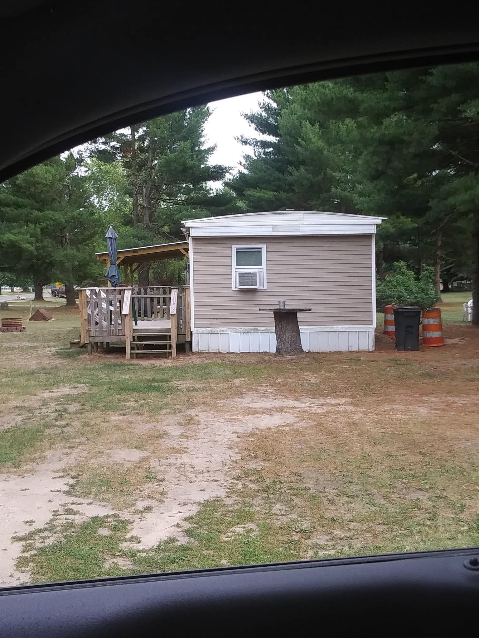 Mobile home with small deck, set on grassy area with trees, viewed from car window.