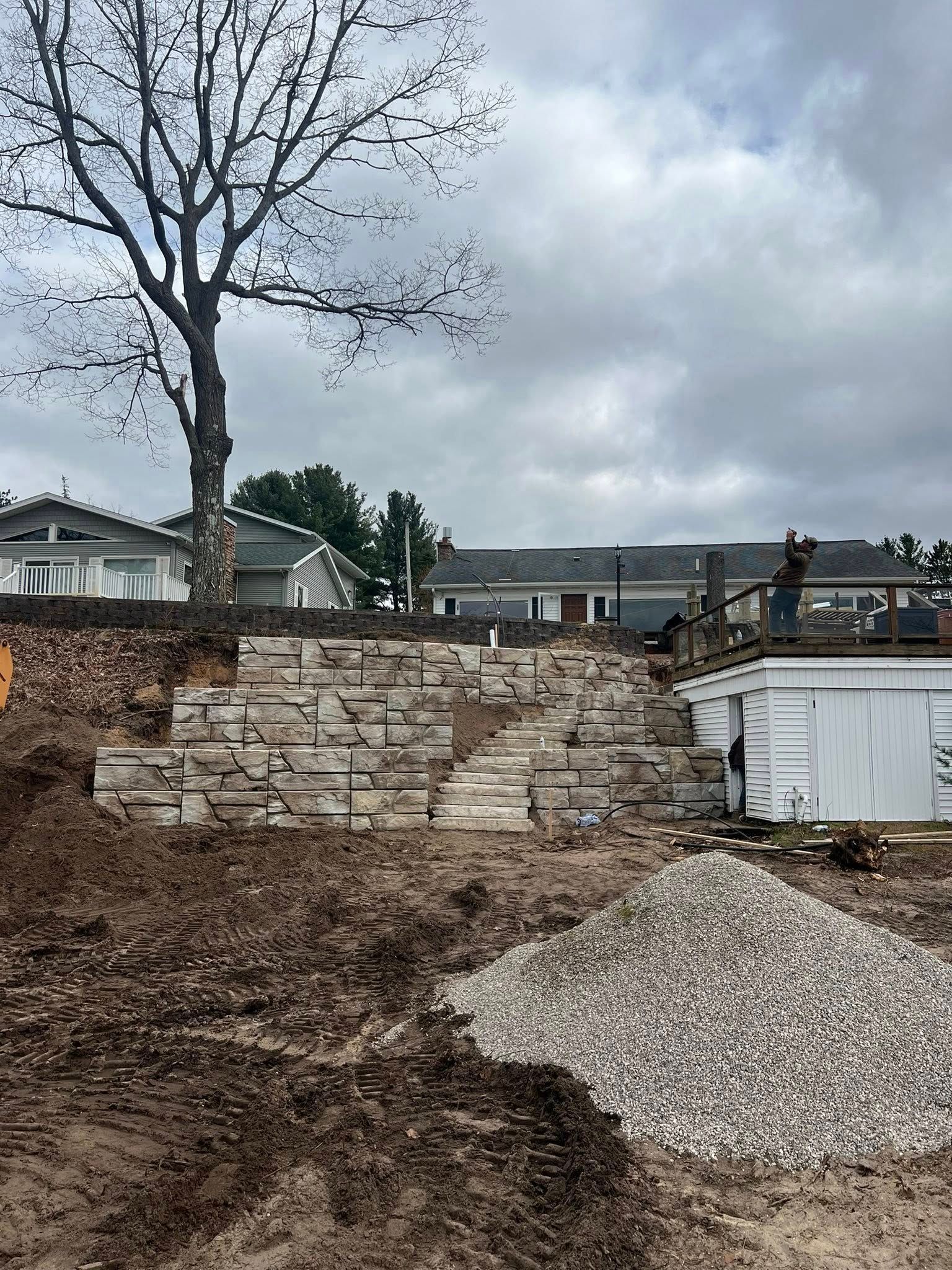 Stone retaining wall under construction next to a pile of gravel with buildings in the background.