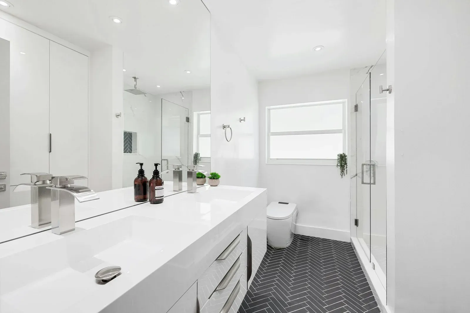 Modern white bathroom with double vanity, dark patterned floor, and glass shower.