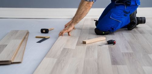 A person installing wood flooring, kneeling, placing a plank. Tools and wood pieces are nearby.
