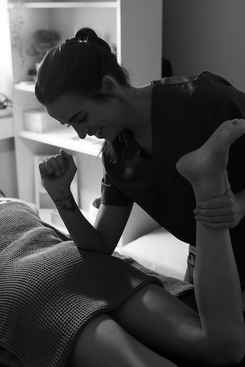 Woman providing massage. Kneeling, smiling, holding patient's leg, arm resting on bed. White shelves in background.