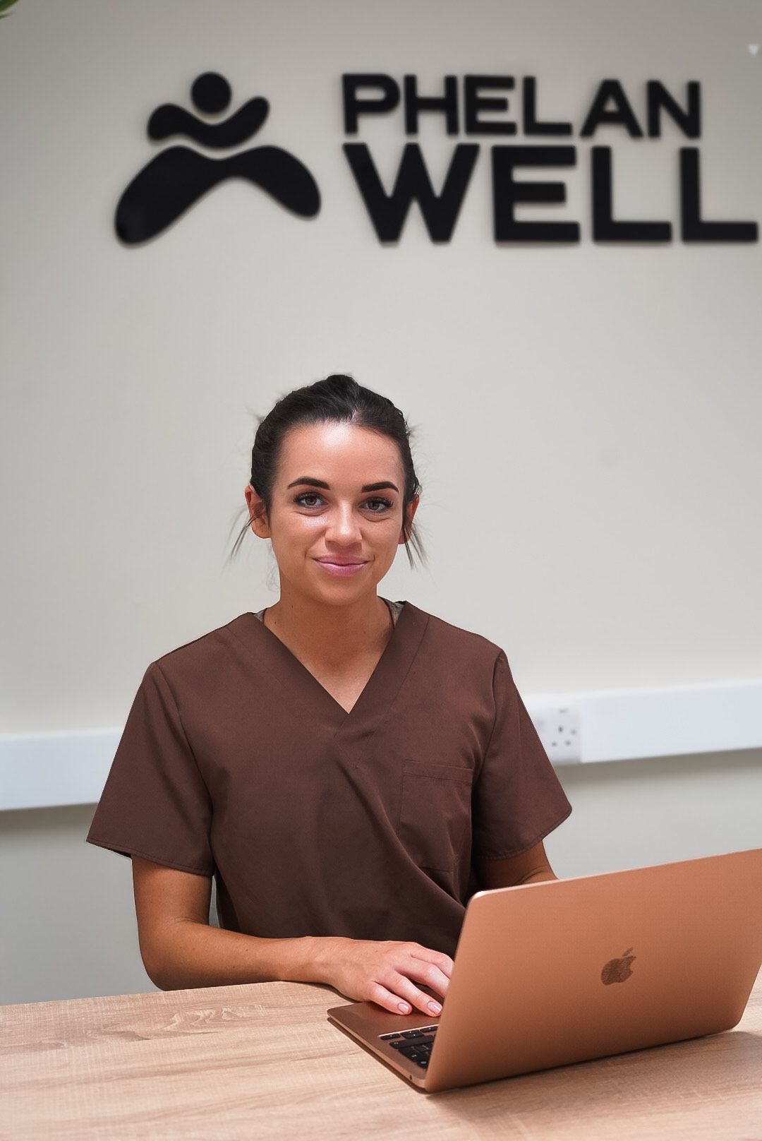 Woman in brown scrubs at desk with laptop, under 