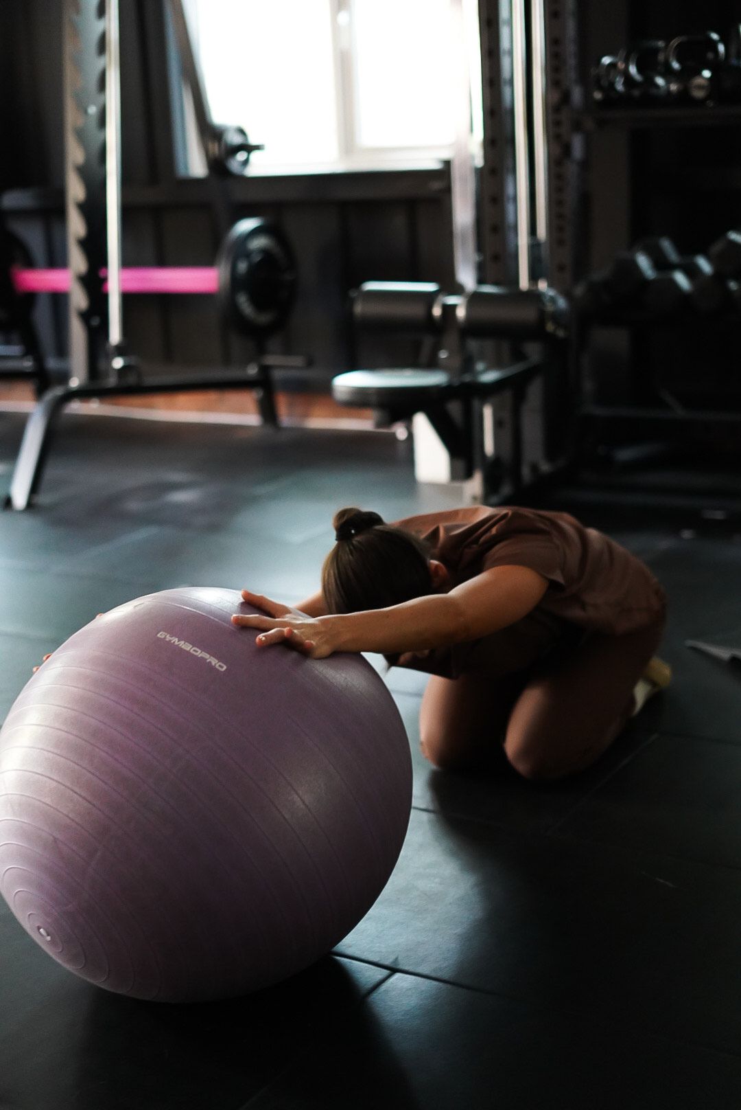 Woman kneeling, stretching over a purple exercise ball in a gym.