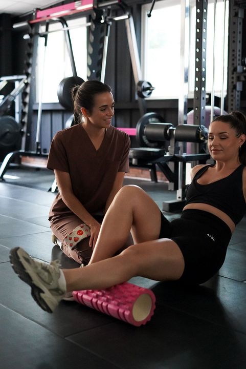Woman using foam roller on leg in gym, another woman watches and smiles.