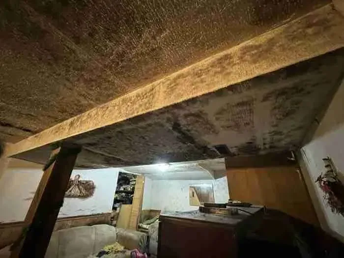 Low-ceiling basement with exposed beams, cluttered furniture, and a bright light in the back room