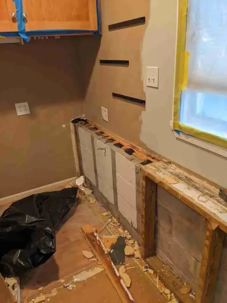 Partially demolished kitchen wall with exposed studs, debris, and a window with taped paper covering