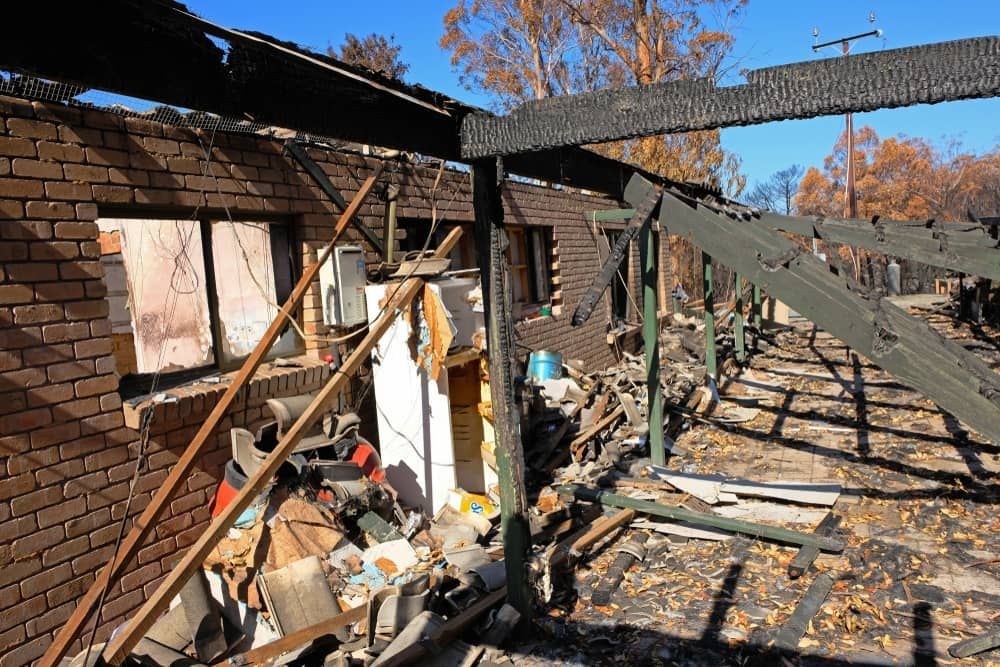Severely burned house interior with collapsed beams, debris, and charred walls under a damaged roof.