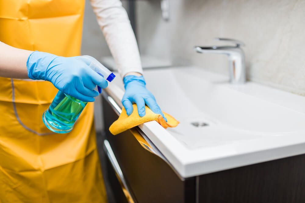 Person in yellow apron cleaning a bathroom sink with spray and cloth