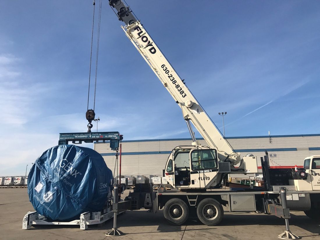 Crane lifting a large blue-wrapped cylindrical load beside a white tanker trailer outdoors.