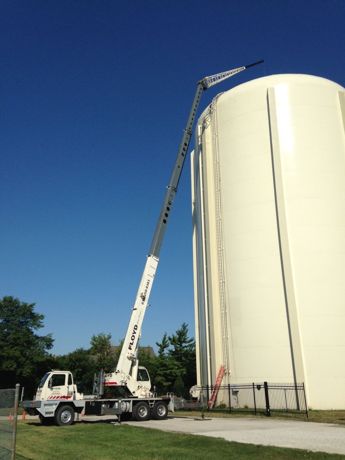Crane truck lifting a long boom beside a large white cylindrical tank under a clear blue sky