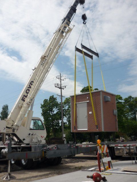 Crane lifting a small brick modular building with yellow straps at a construction site