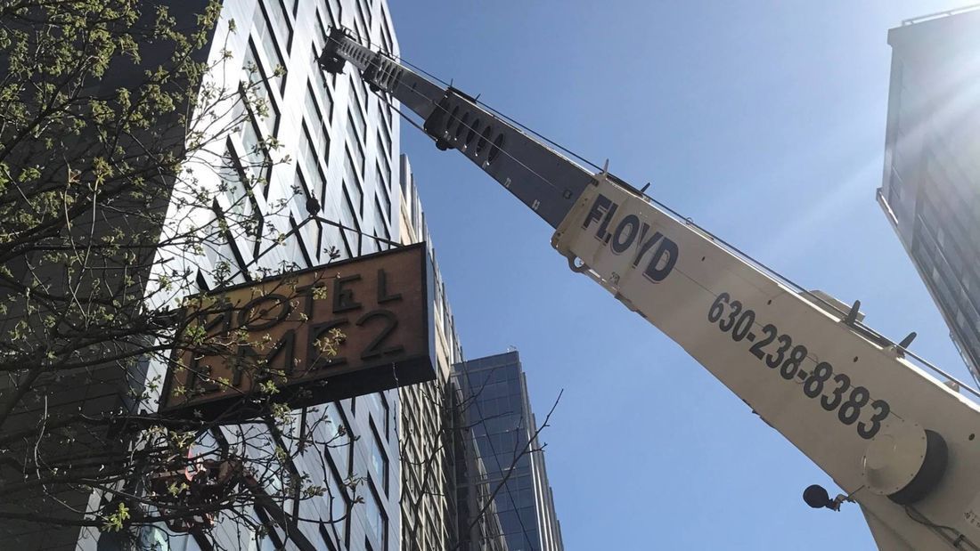 Crane lifting a hotel sign beside a tall building under a bright sky