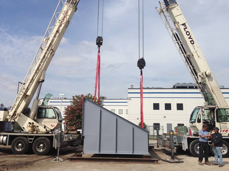 Two cranes lifting a large gray wall panel at a construction site