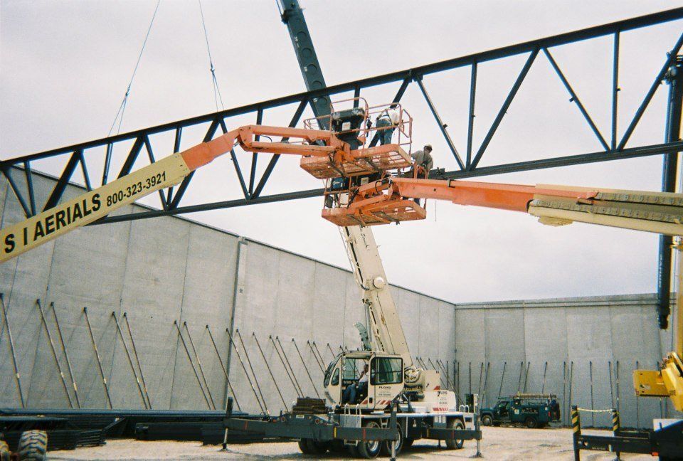 Two orange boom lifts reaching up to a steel truss inside a large industrial building frame