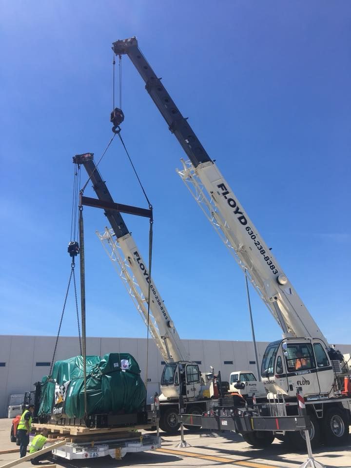 Two cranes lifting a large green wrapped load in an outdoor industrial yard under a blue sky