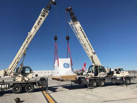 Two cranes lifting a large white GE load on a flatbed truck under a blue sky. Two cranes lifting a large white GE load on a flatbed truck under a blue sky.