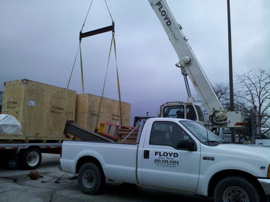 Crane lifting a large crate onto a white Floyd pickup truck at a work site.