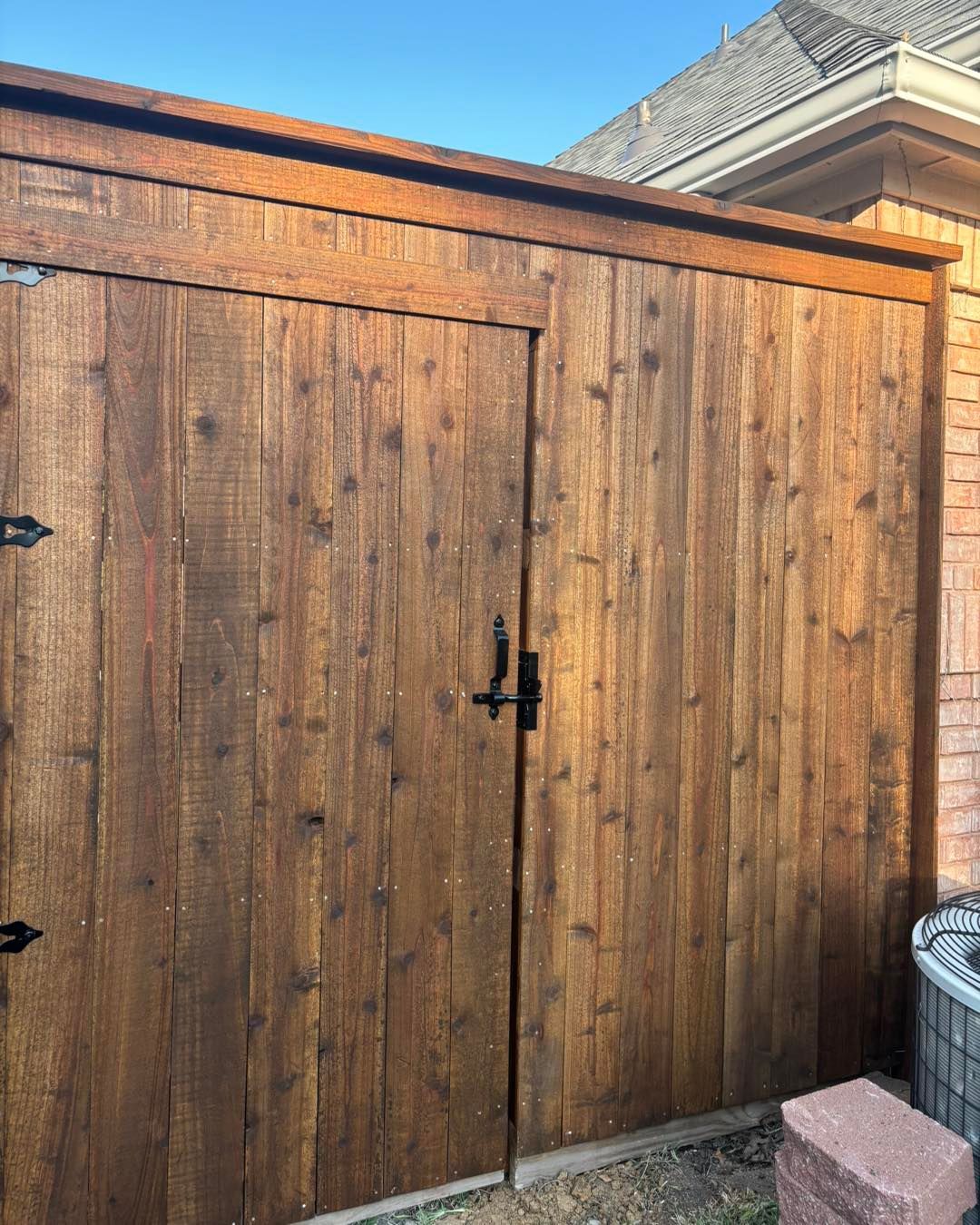 Wooden shed doors, stained brown, with black hardware, outside near a brick wall.