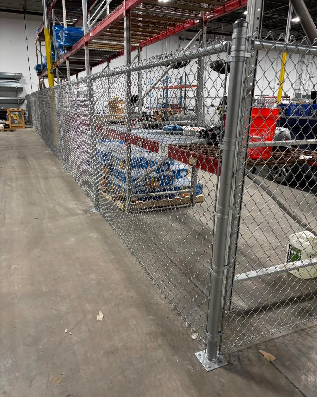 Chain-link fence inside a warehouse, separating stored pallets and items. Metal supports and overhead shelving are visible.