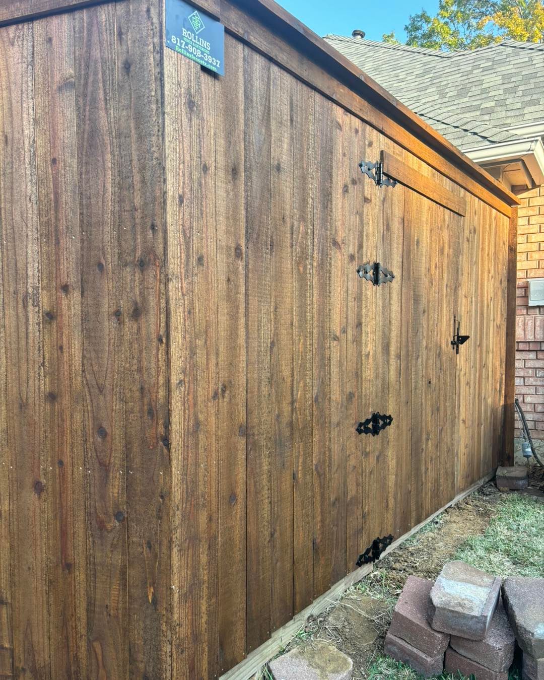 Wooden shed with brown stain and black hinges next to a brick house and green grass.