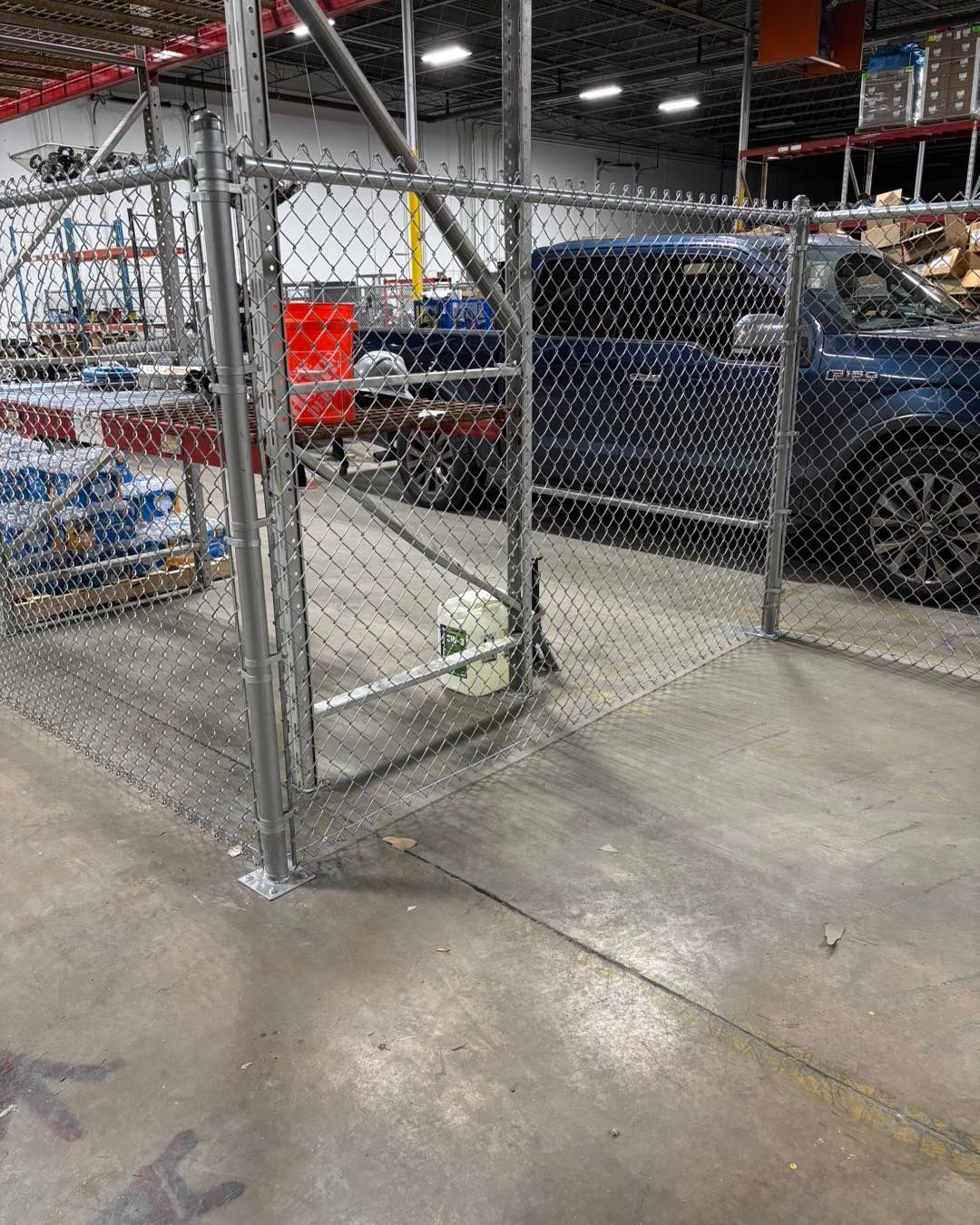 Chain-link fence with a closed gate; a car and a red cart sit behind the fence inside a warehouse.