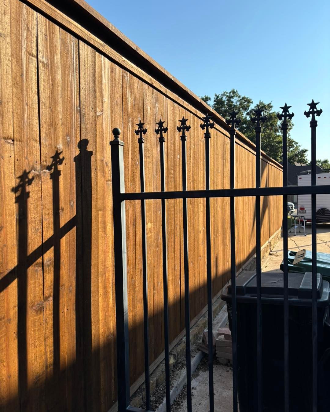 A dark metal fence with ornate tops casts shadows on a wooden fence in sunlight.