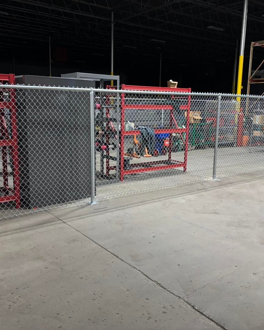 Chain-link fence encloses a warehouse area with red racks holding items on gravel ground.