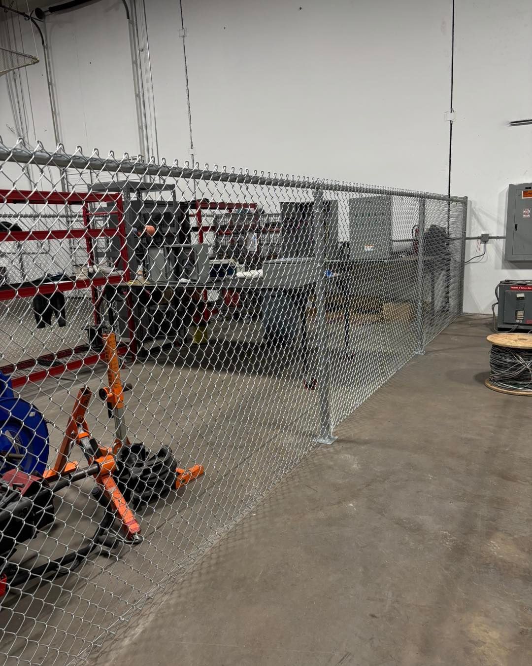 Chain-link fence within an industrial space, enclosing equipment against a white wall and concrete floor.