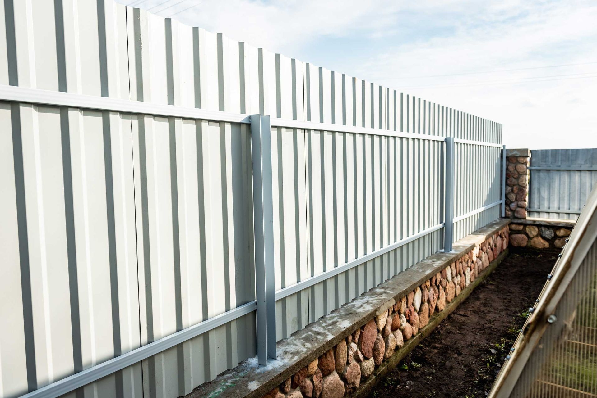 A light gray corrugated metal privacy fence installed over a stone foundation wall against a bright, cloudy sky.