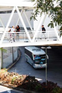 Ônibus passando sob uma passarela branca. Pessoas caminhando sobre a passarela.