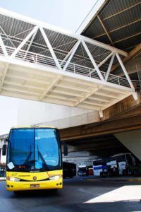 Ônibus amarelo passando por baixo de uma passarela branca em uma rodoviária. Outros ônibus são visíveis.