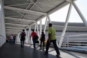 Pessoas caminhando por uma passarela coberta com estrutura metálica. Luz solar intensa.