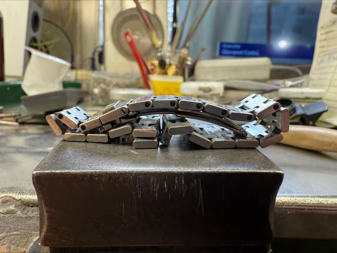 In un'officina disordinata, un cinturino metallico per orologio giace piegato in una forma curva su un blocco rettangolare di acciaio da gioielliere.