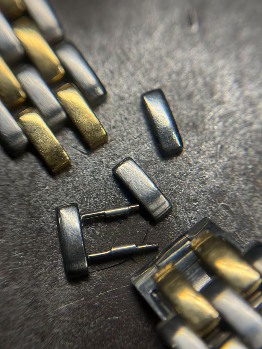 Un cinturino di orologio in metallo rotto, con maglie allentate color argento e due sottili perni di collegamento, giace su una superficie scura e ruvida.