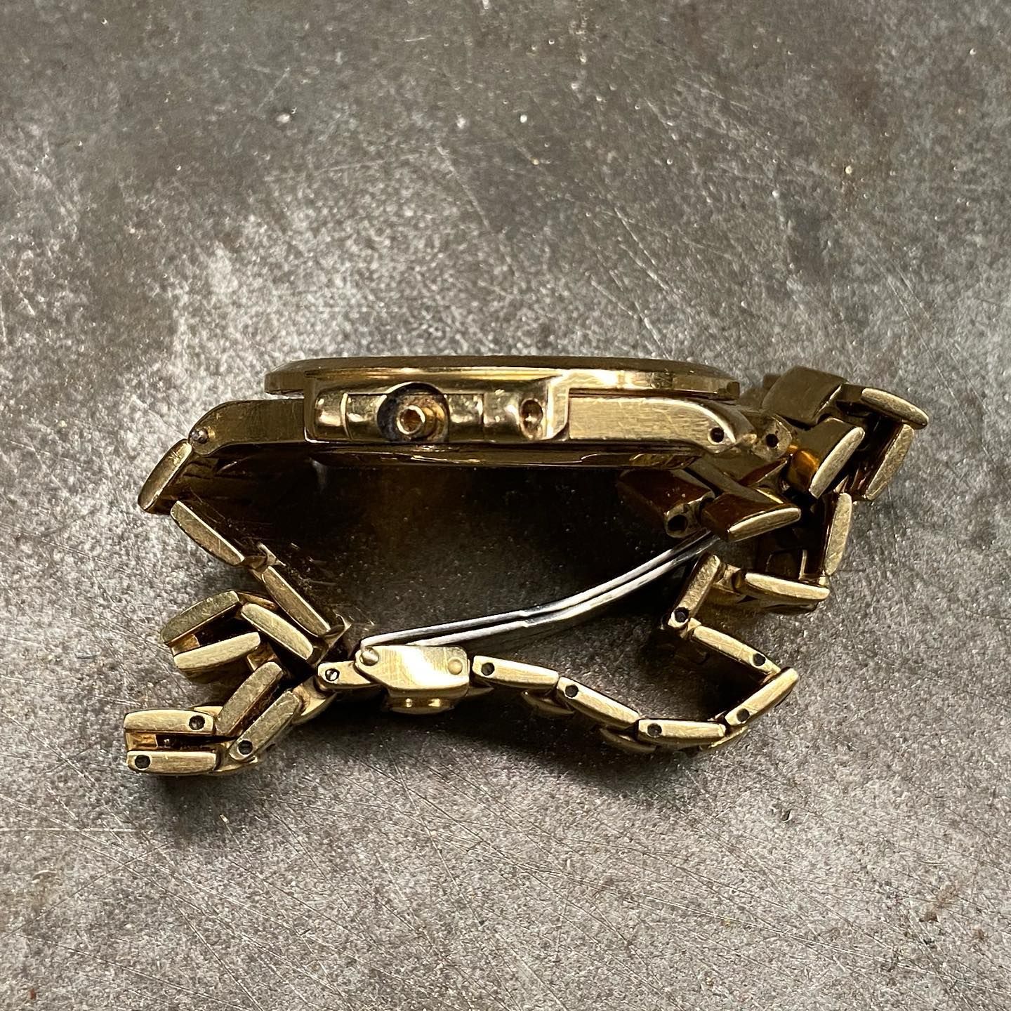 Vista laterale di un orologio da polso in metallo color oro con bracciale a maglie piegate, appoggiato su una superficie grigia e ruvida.