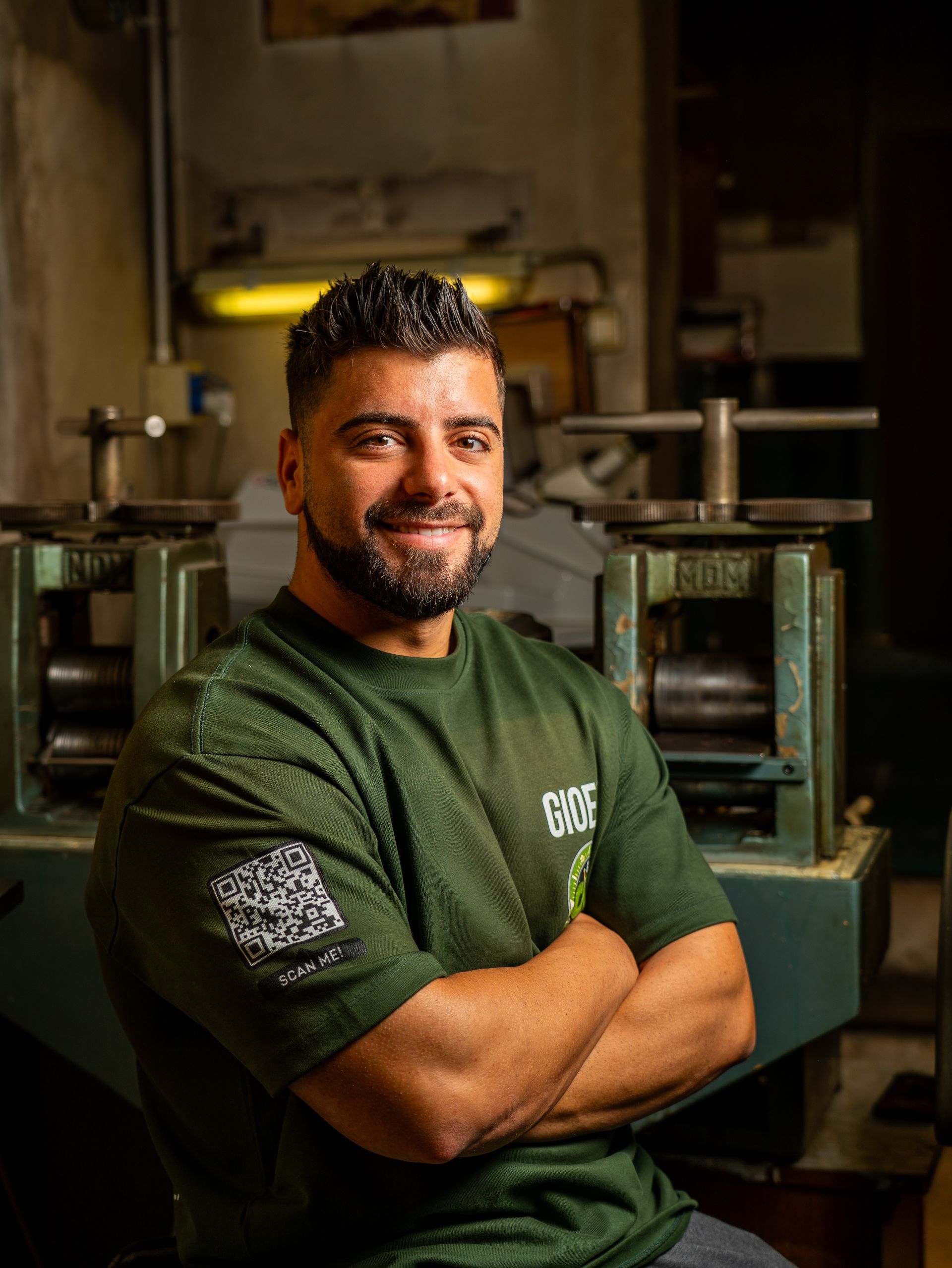 In un'officina con attrezzature metalliche, una persona con la barba e i capelli scuri sorride a braccia conserte, indossando una maglietta verde.