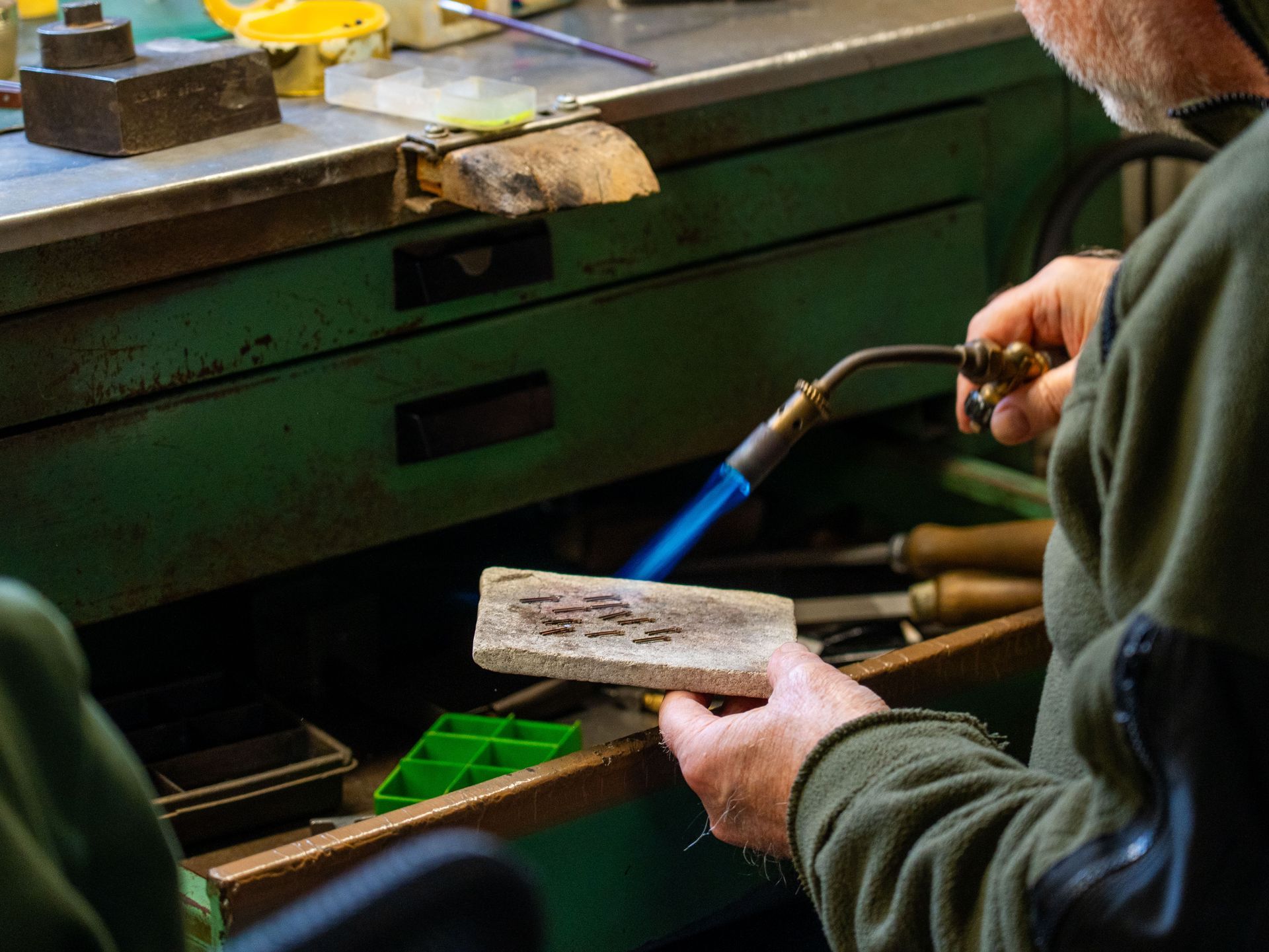Un gioielliere utilizza una torcia a mano per applicare una fiamma blu a un pezzo di metallo appoggiato su un blocco di saldatura su un banco da lavoro.
