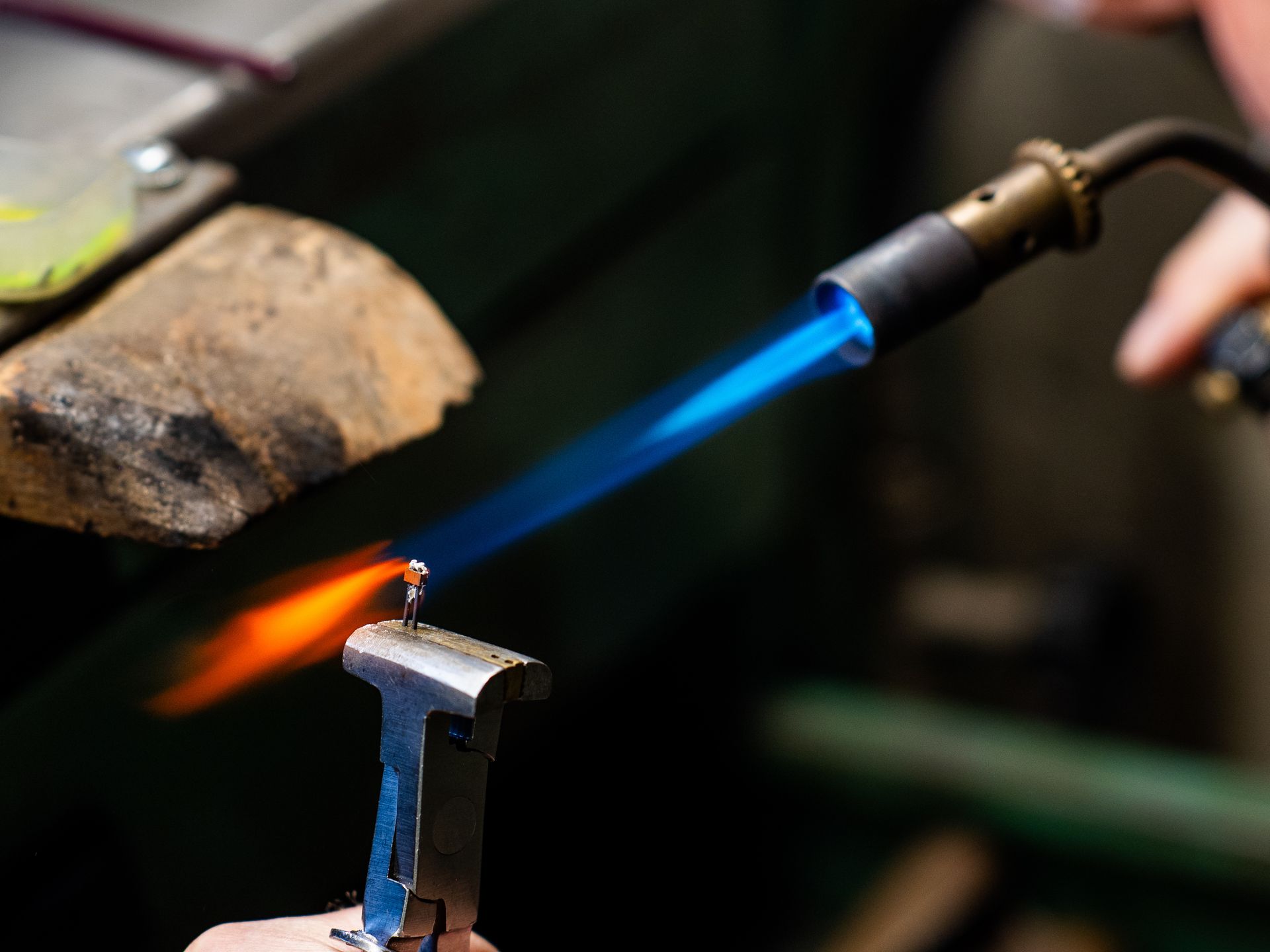 Un gioielliere utilizza una torcia portatile per applicare una fiamma blu brillante a un piccolo pezzo di metallo tenuto in una morsa.
