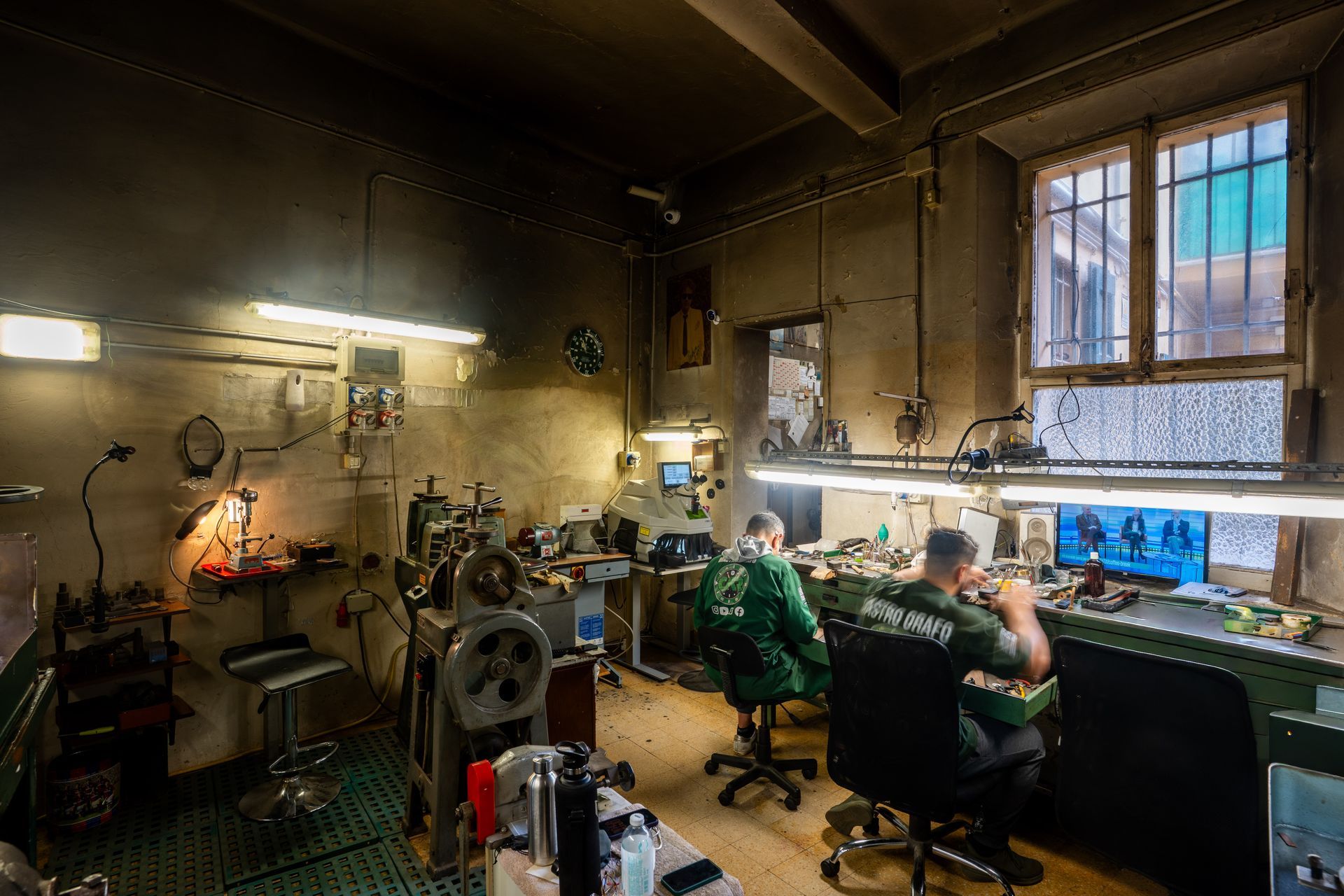 Due persone in abiti da lavoro scuri siedono alle postazioni di lavoro in un'officina industriale poco illuminata, riparando attrezzature alla luce delle lampade da banco.