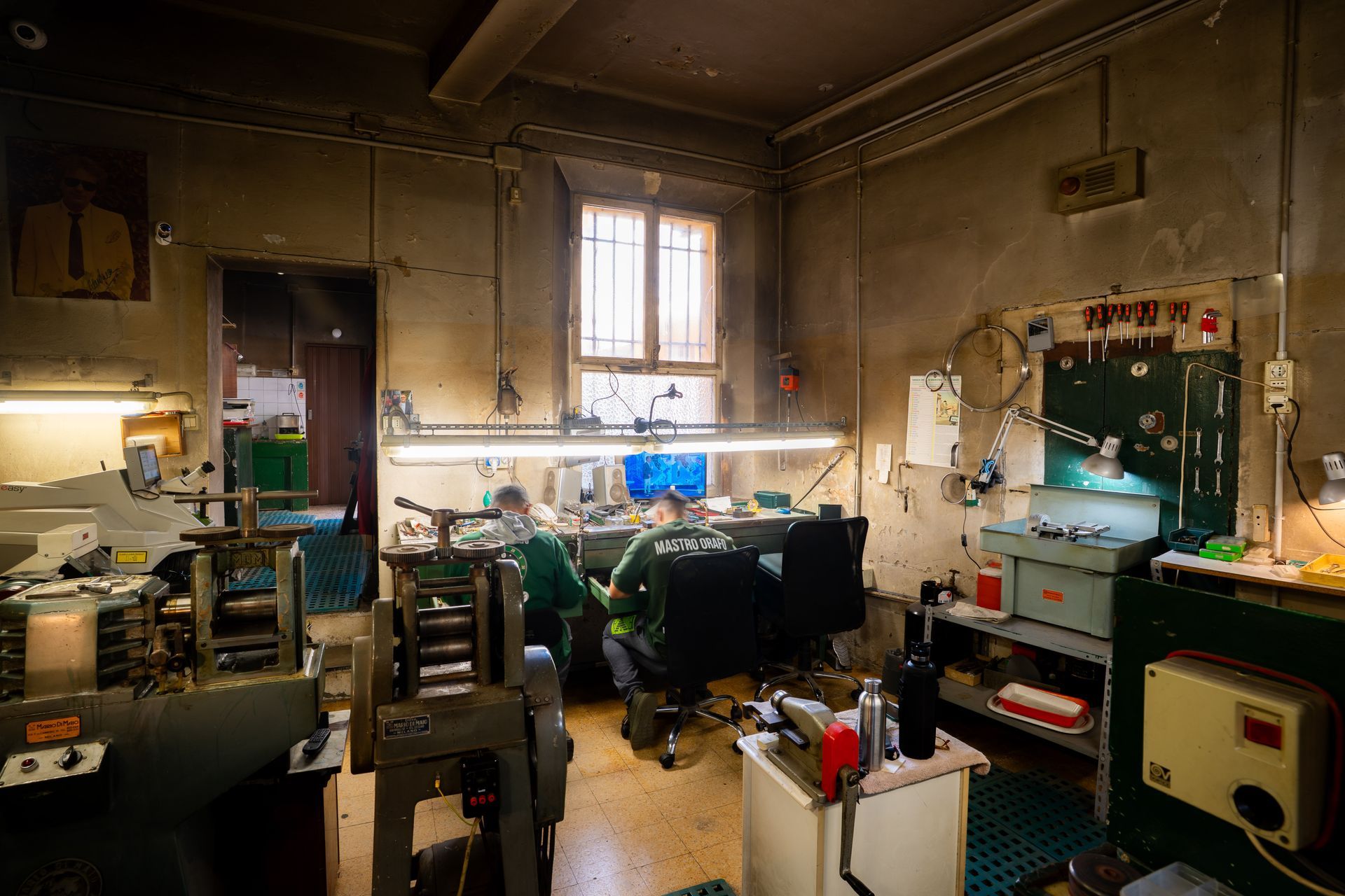 In un'officina rustica in stile industriale, due artigiani lavorano su banchi ingombri di attrezzi sotto luci intense.