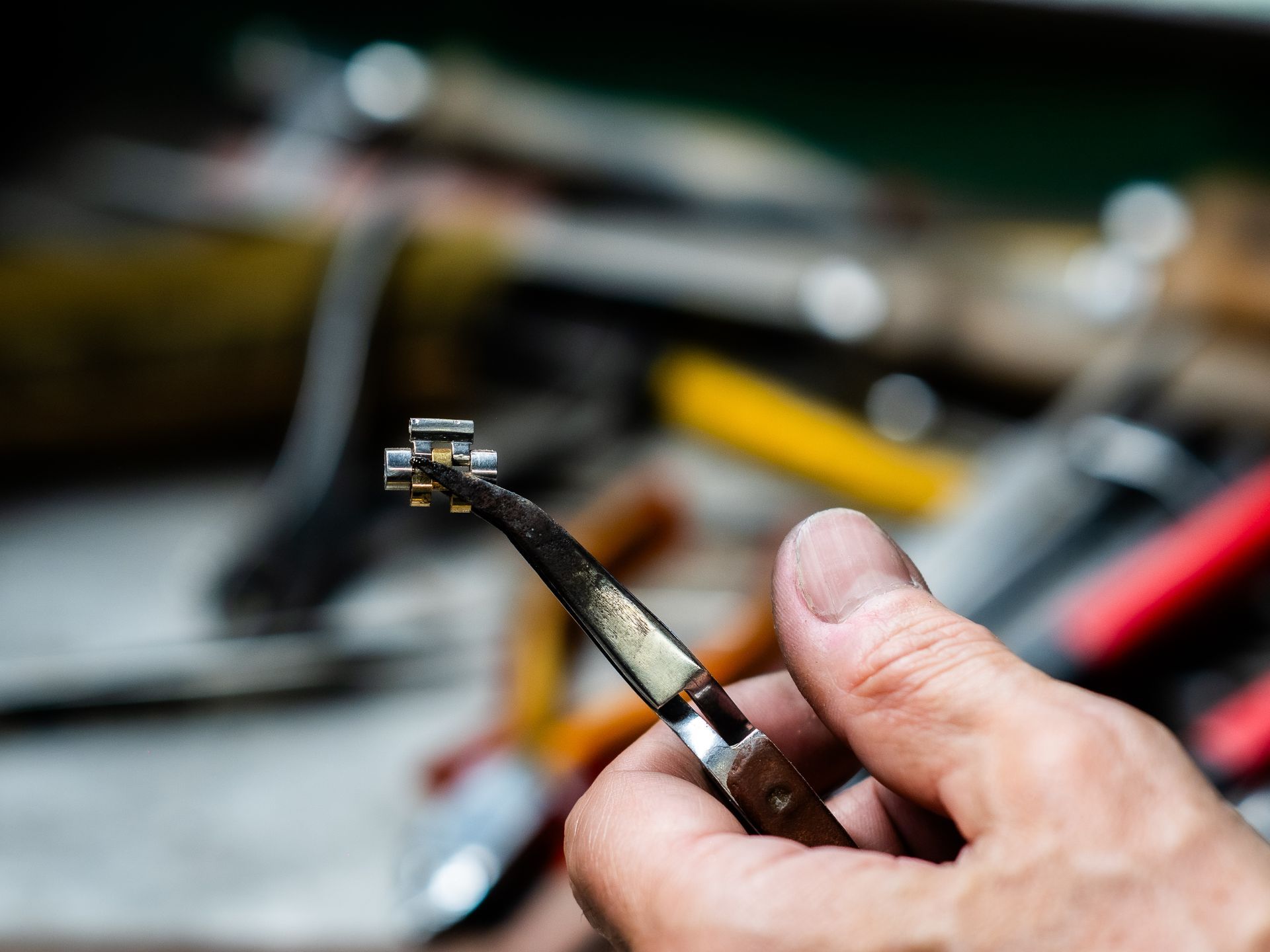 Una mano stringe con una pinza di precisione un piccolo componente metallico di un orologio sopra un banco da lavoro pieno di vari attrezzi.