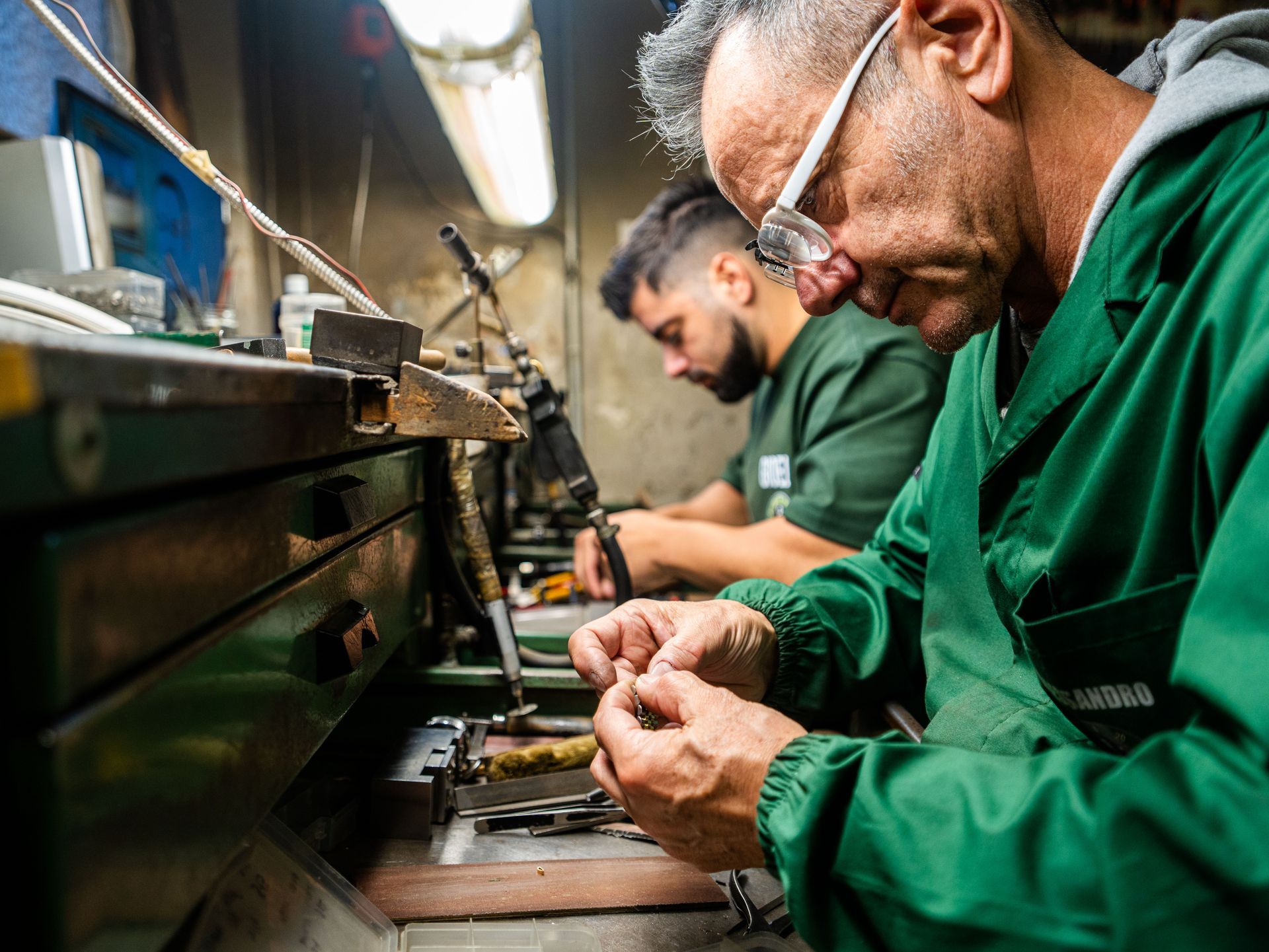 Due persone con indosso camici da lavoro verdi sono sedute a un banco da lavoro in un'officina, concentrate su un lavoro manuale di precisione.