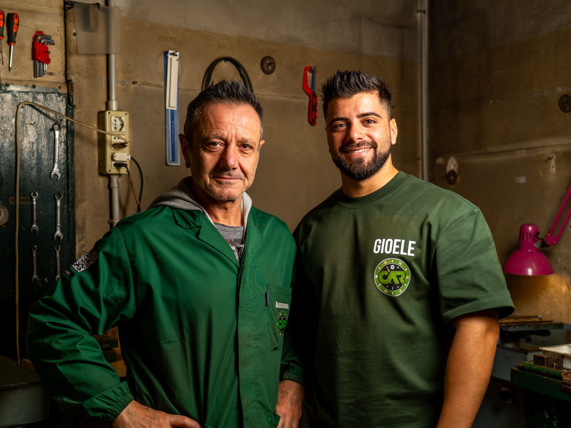Due uomini in abiti da lavoro verdi sono in piedi in un'officina, in posa davanti ad attrezzi appesi a una parete.