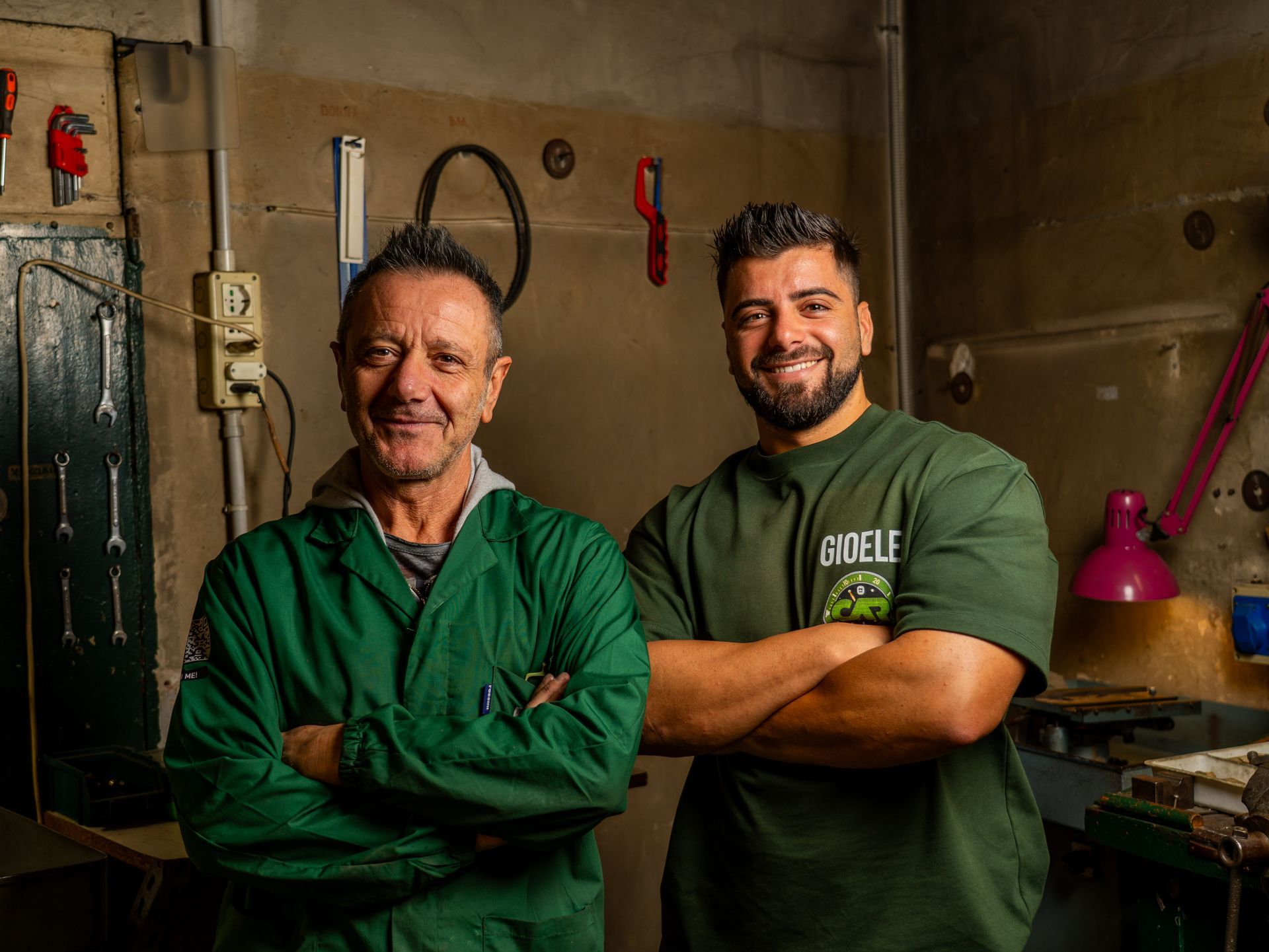 Due uomini con camicie verdi sono in piedi in un'officina con le braccia incrociate e sorridono alla telecamera.