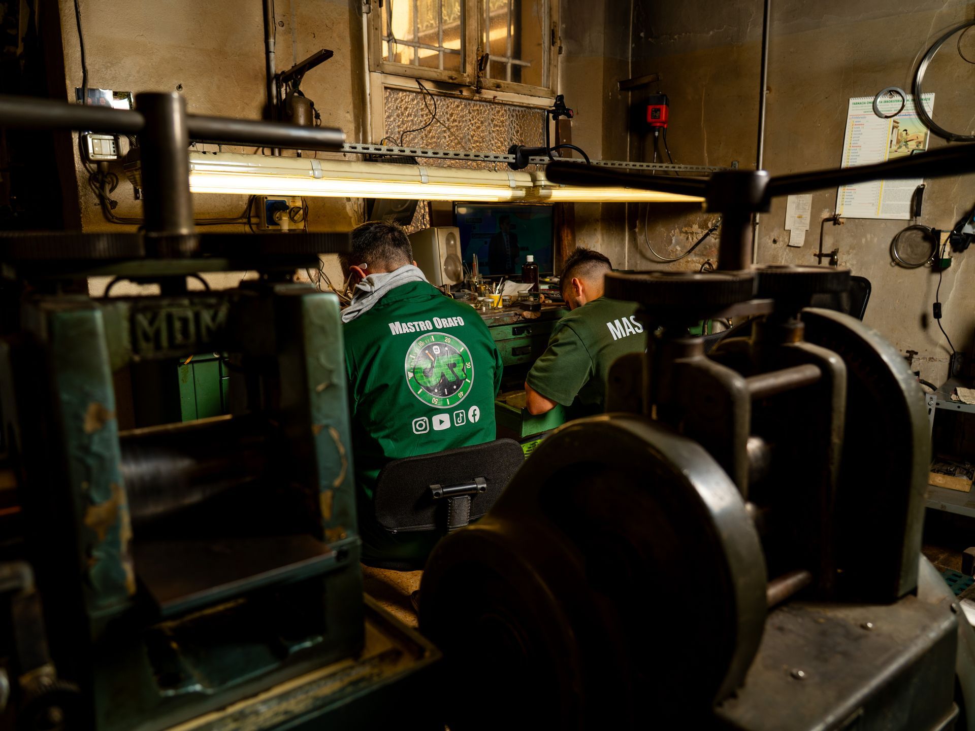 Due persone con indosso camicie verdi lavorano a delle macchine per la laminazione dei metalli in un'officina industriale scarsamente illuminata.