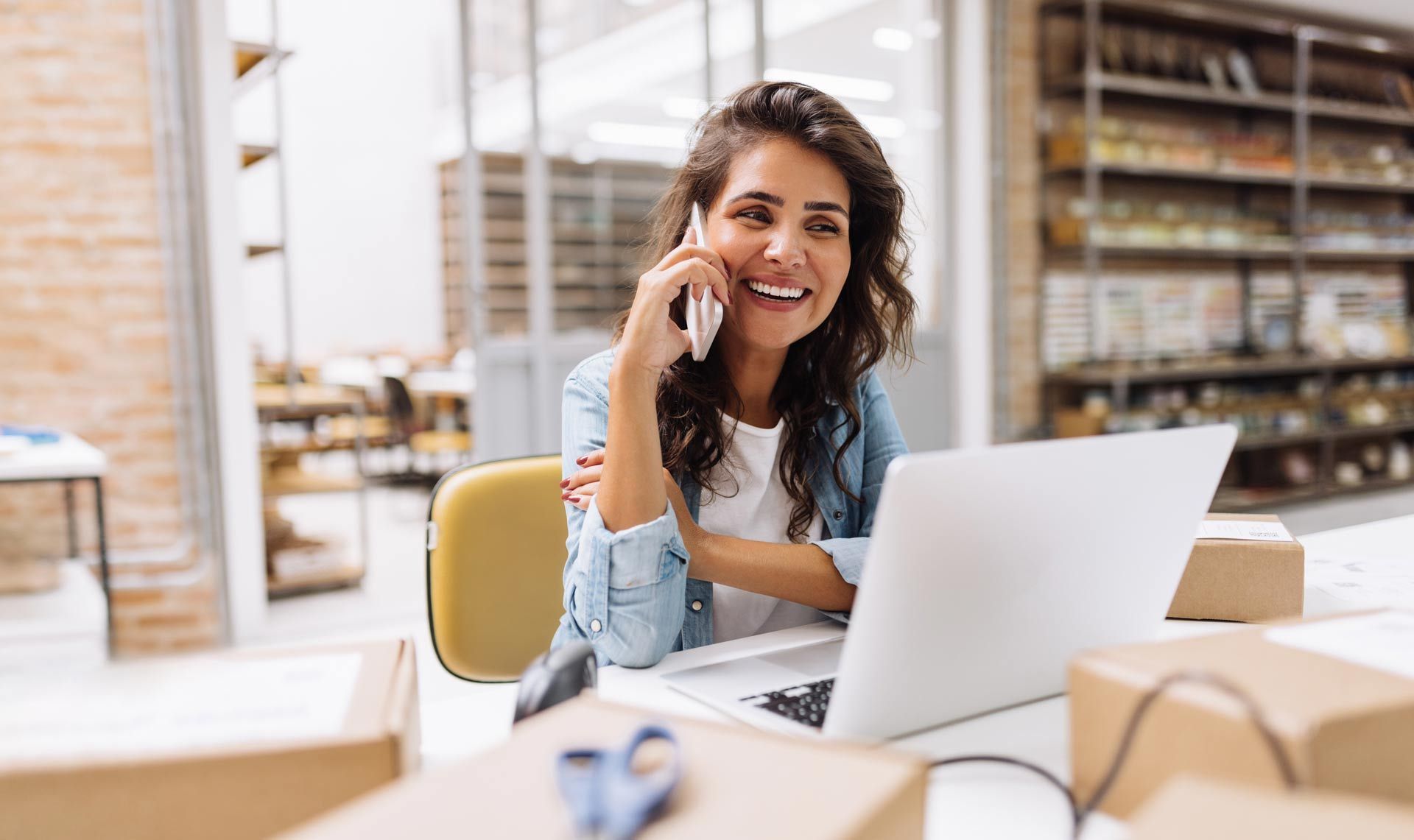 A woman is sitting at a desk with a laptop and talking on a cell phone.