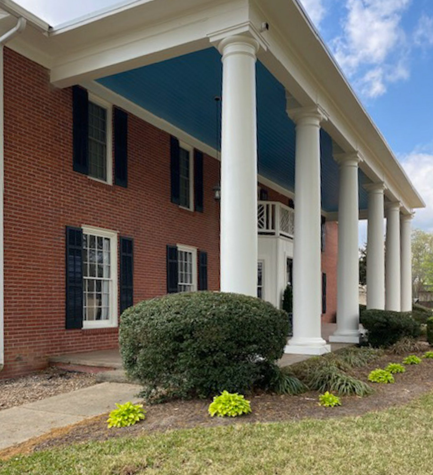 Brick building with white columns, black shutters, and trimmed shrubs along the entrance.