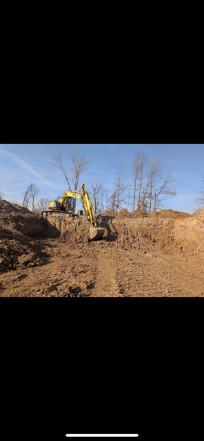 A yellow excavator digs into a brown dirt hill under a blue sky, trees in the background.